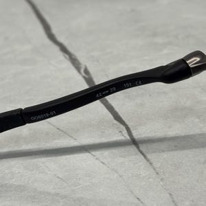 May include: Close-up of a black eyeglass frame with the text "OO6019-01", "42 29", and "151 CE" printed on the arm. The frame is made of a matte black material.