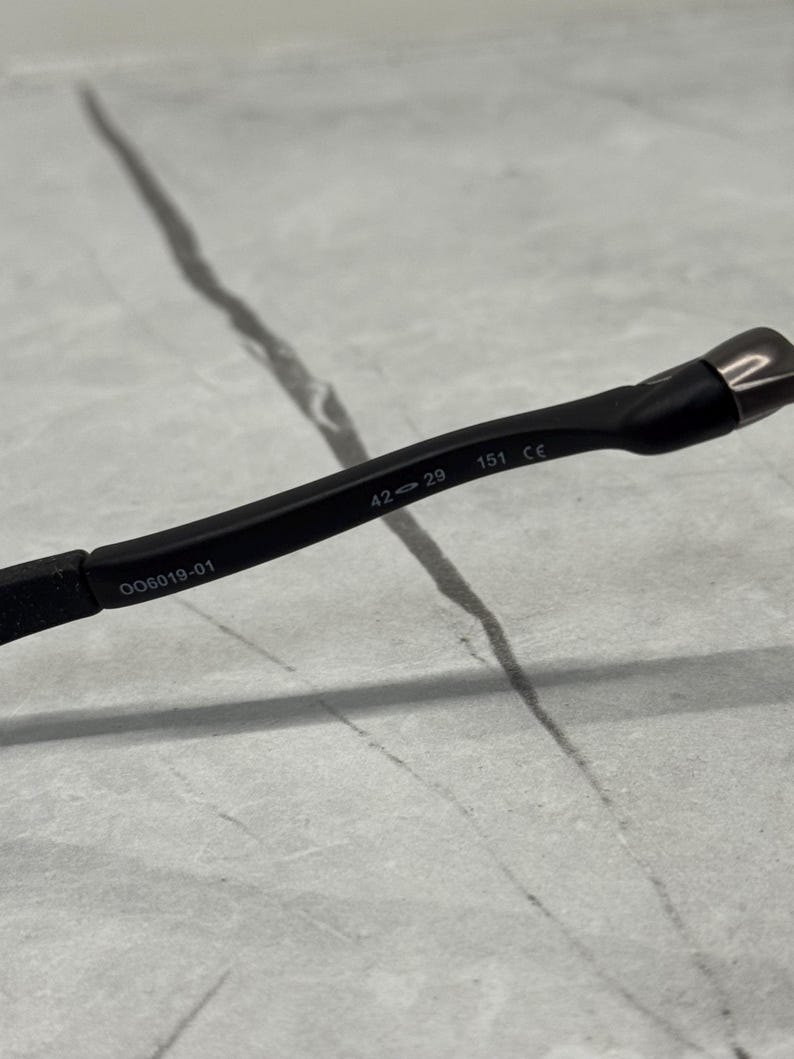 May include: Close-up of a black eyeglass frame with the text "OO6019-01", "42 29", and "151 CE" printed on the arm. The frame is made of a matte black material.