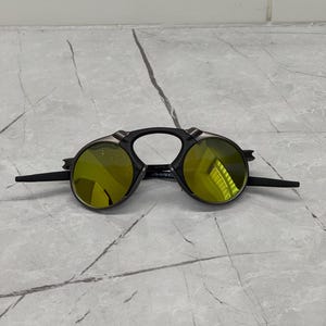 May include: Black sunglasses with round, yellow-tinted lenses. The sunglasses have a unique design with a black frame and extended temples. The lenses reflect light, and the sunglasses are set against a gray marble surface.