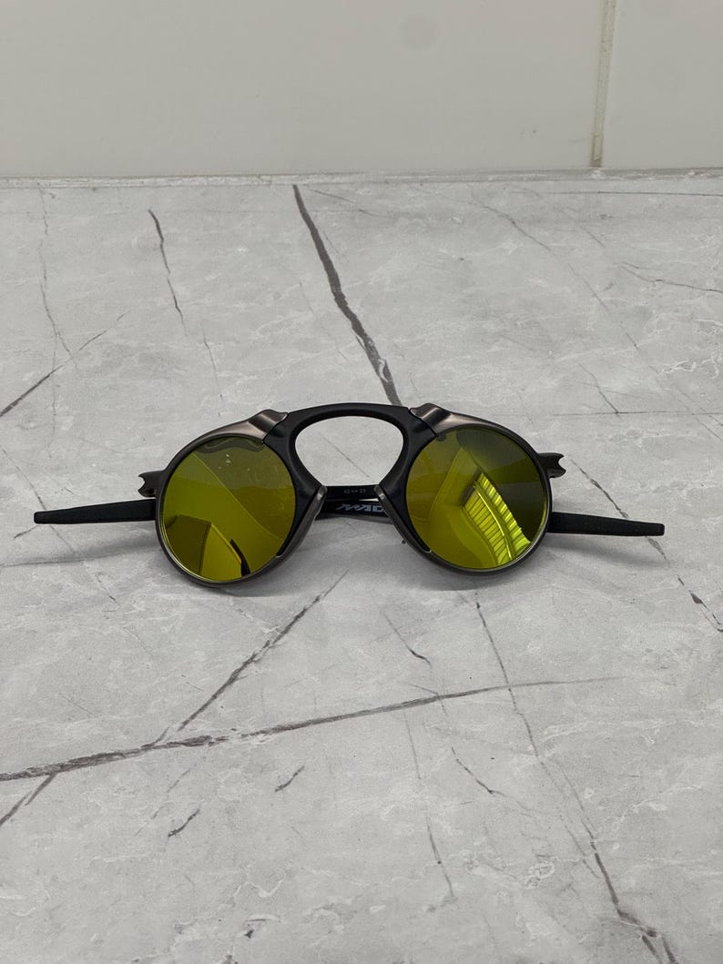 May include: Black sunglasses with round, yellow-tinted lenses. The sunglasses have a unique design with a black frame and extended temples. The lenses reflect light, and the sunglasses are set against a gray marble surface.