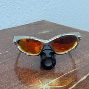 Puede incluir: Gafas de sol con montura plateada y lentes naranjas y un accesorio cil&iacute;ndrico negro. Las gafas de sol tienen un dise&ntilde;o curvo y est&aacute;n sobre una superficie de madera. Las lentes son reflectantes.