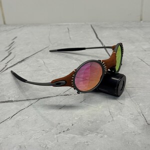 以下が含まれることがあります： 丸いレンズのサングラス。フレームの周りに茶色の革のアクセントがあります。レンズはピンクとオレンジのグラデーションです。テンプルは黒くて細いです。サングラスは黒い円筒形の物体の上に置かれています。