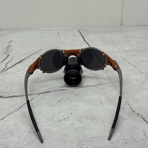 May include: Oval sunglasses with dark lenses and a brown leather bridge. The frames are a gunmetal color with black temple tips. The sunglasses have a unique, vintage-inspired design.