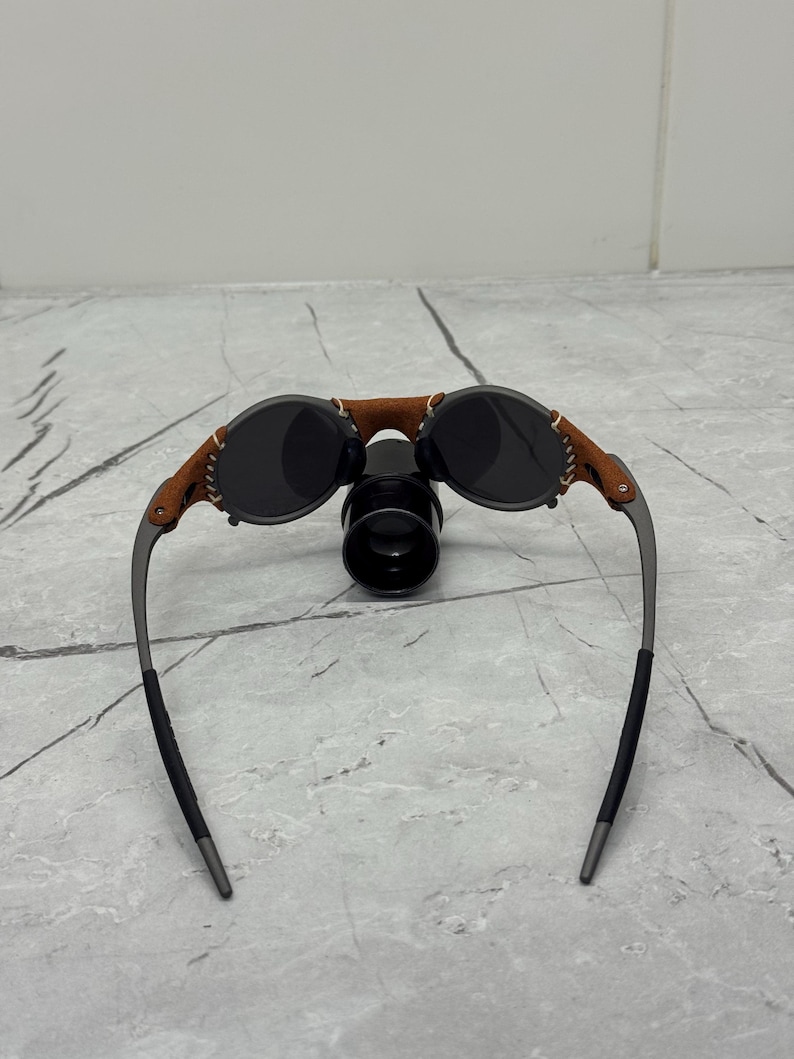 May include: Oval sunglasses with dark lenses and a brown leather bridge. The frames are a gunmetal color with black temple tips. The sunglasses have a unique, vintage-inspired design.