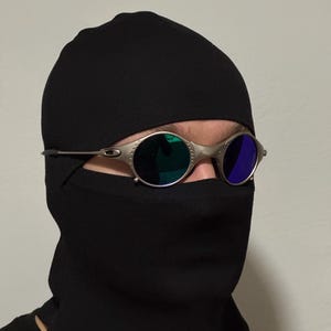 Może przedstawiać: Srebrne okulary przeciwsłoneczne z zielonymi i niebieskimi soczewkami noszone na czarnej kominiarce. Kominiarka zakrywa głowę i szyję, pozostawiając widoczne tylko oczy. Okulary przeciwsłoneczne mają okrągły kształt.