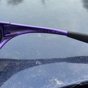 May include: Close-up of purple sunglasses with black earpieces. The text "CE MADE IN U.S.A." is printed on the temple arm. The sunglasses are likely designed for outdoor activities.