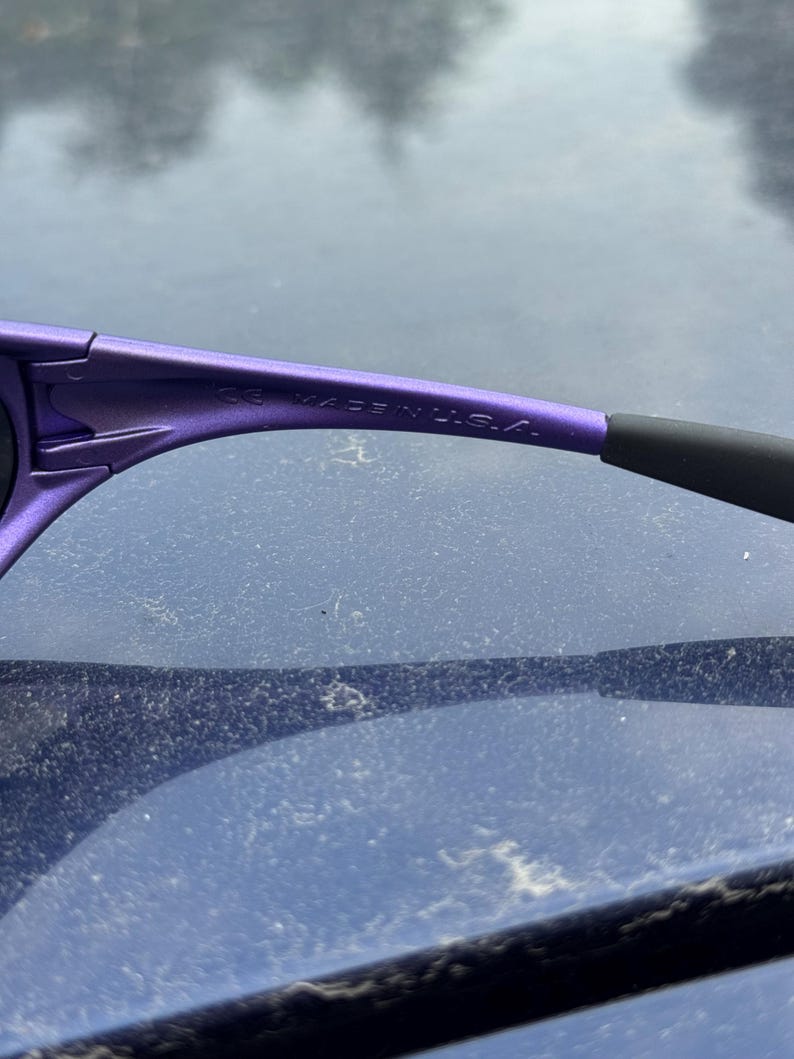 May include: Close-up of purple sunglasses with black earpieces. The text "CE MADE IN U.S.A." is printed on the temple arm. The sunglasses are likely designed for outdoor activities.