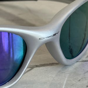 May include: Close-up of silver sunglasses with blue and purple lenses. The brand name "Oakley" is printed on the frame. The sunglasses have a sleek, modern design.
