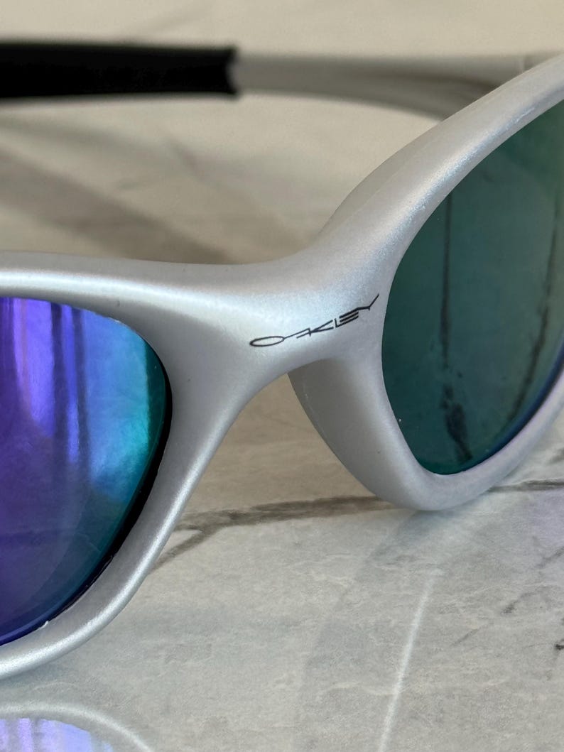 May include: Close-up of silver sunglasses with blue and purple lenses. The brand name "Oakley" is printed on the frame. The sunglasses have a sleek, modern design.