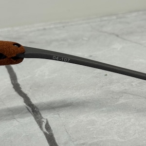 May include: Close-up of a portion of eyeglasses. The frame is dark gray with the text "04-107" printed on the arm. The temple tips are covered in a brown, suede-like material.