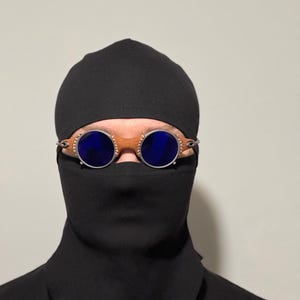 May include: Round blue-lensed goggles with a brown leather bridge and silver-toned metal frames. The goggles are worn over a black balaclava and a black garment, creating a mysterious look. The background is a plain, light color.