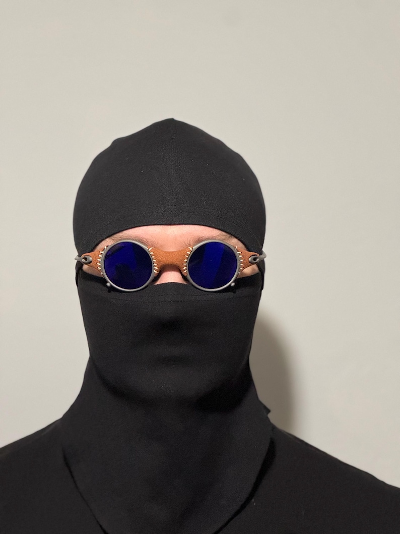 May include: Round blue-lensed goggles with a brown leather bridge and silver-toned metal frames. The goggles are worn over a black balaclava and a black garment, creating a mysterious look. The background is a plain, light color.