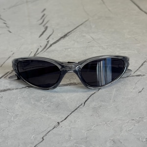 May include: Sport sunglasses with a gray frame and dark lenses. The sunglasses have a curved design and a sleek, modern aesthetic. The lenses are reflective, and the frame is a translucent gray color.