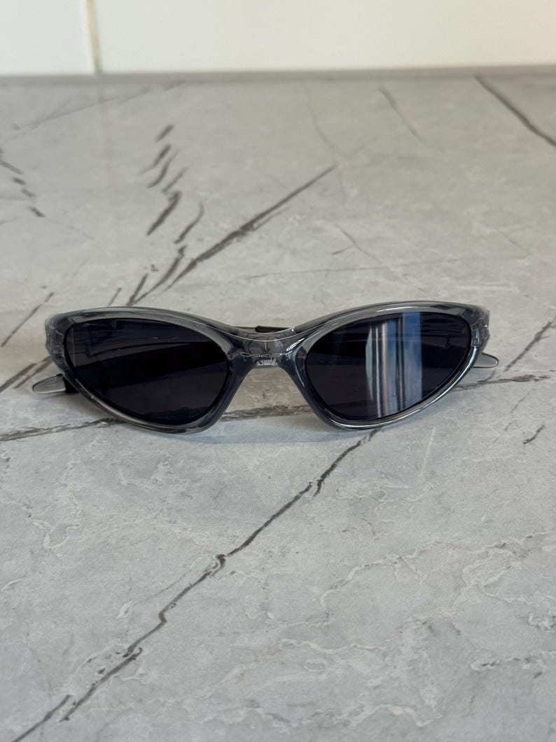 May include: Sport sunglasses with a gray frame and dark lenses. The sunglasses have a curved design and a sleek, modern aesthetic. The lenses are reflective, and the frame is a translucent gray color.