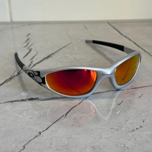 May include: Silver-framed sunglasses with red-orange lenses. The sunglasses have a curved design with black accents on the temples and arms. The lenses reflect light, and the overall style is sporty and modern.