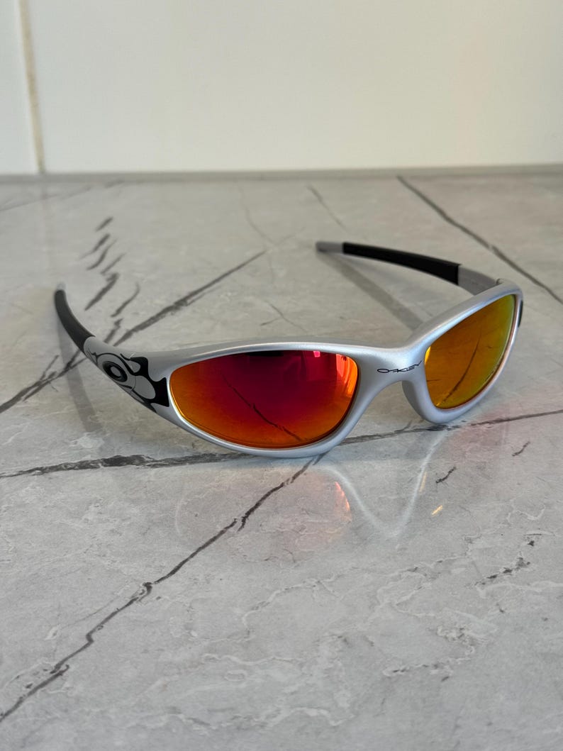 May include: Silver-framed sunglasses with red-orange lenses. The sunglasses have a curved design with black accents on the temples and arms. The lenses reflect light, and the overall style is sporty and modern.