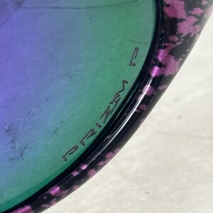 May include: Close-up of sunglasses with a black frame and purple and pink speckled accents. The lenses have a gradient of green and purple. The word "PRIZM" is printed on the lens.