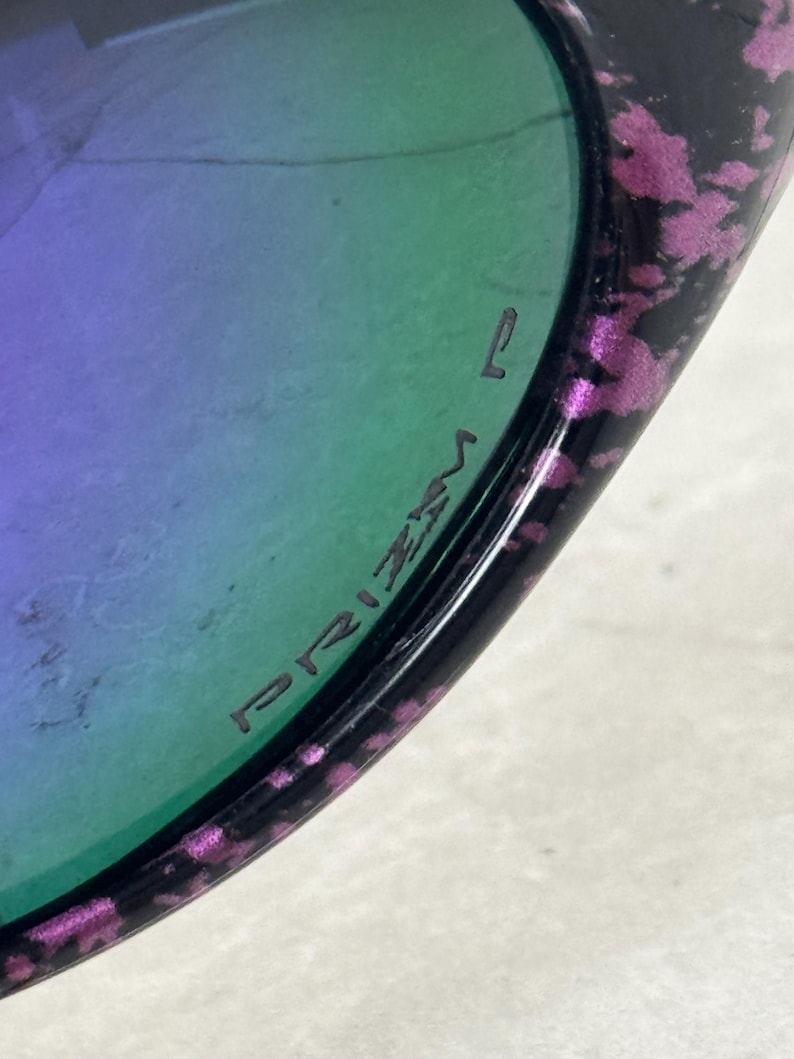May include: Close-up of sunglasses with a black frame and purple and pink speckled accents. The lenses have a gradient of green and purple. The word "PRIZM" is printed on the lens.