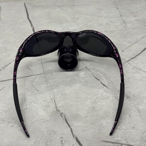 May include: Black sunglasses with a black nose bridge and black lenses. The frames are black with a pink and purple speckled pattern. The arms are black with a pink speckled pattern and have black rubber ear pieces.