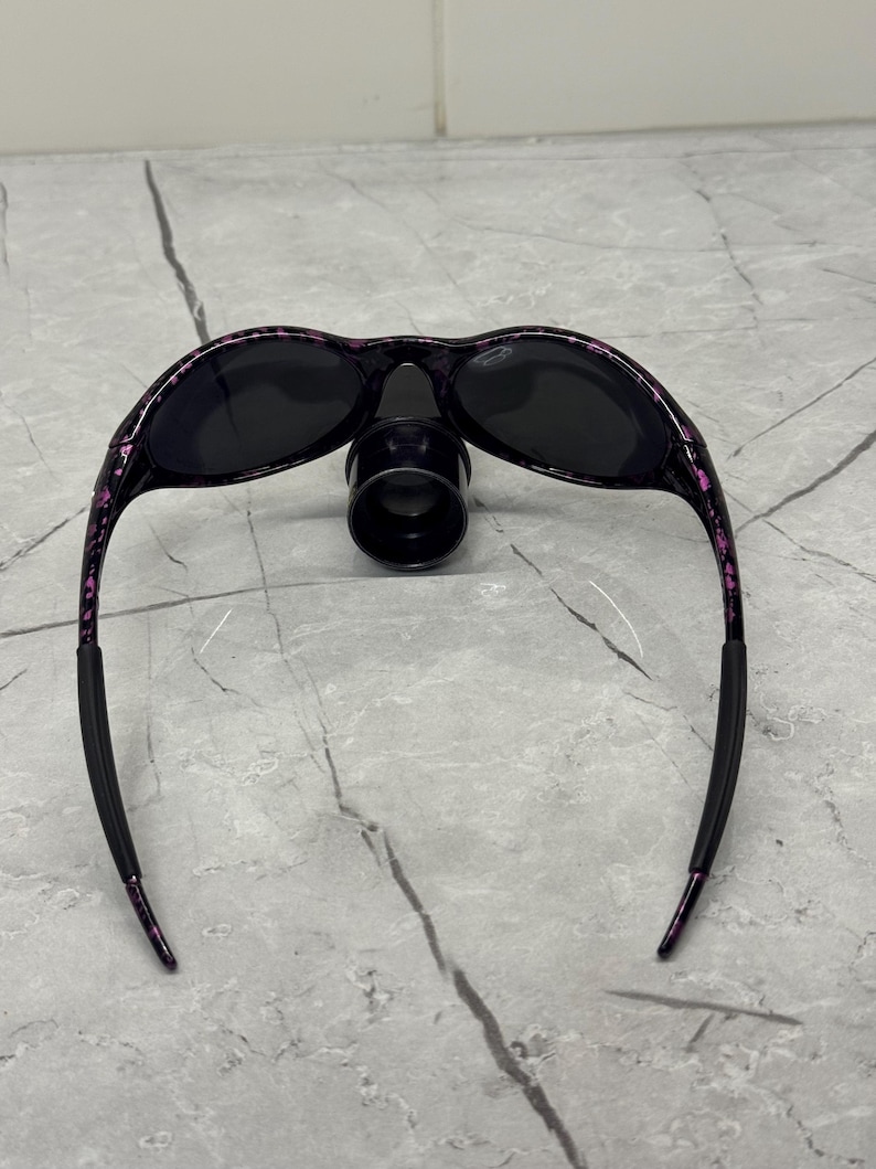 May include: Black sunglasses with a black nose bridge and black lenses. The frames are black with a pink and purple speckled pattern. The arms are black with a pink speckled pattern and have black rubber ear pieces.