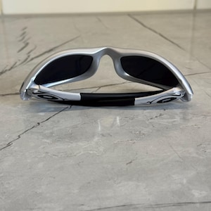 May include: Silver-framed sunglasses with black lenses. The sunglasses are folded, revealing the inner side of the frame and the black rubber nose bridge. The temples feature a white graphic design. The sunglasses are resting on a gray surface.