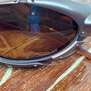 Puede incluir: Primer plano de unas gafas de sol marrones con montura gris oscuro. Las lentes son de color marr&oacute;n degradado. La montura tiene un dise&ntilde;o &uacute;nico con remaches visibles y una forma curva. Las gafas de sol est&aacute;n sobre una superficie de madera.