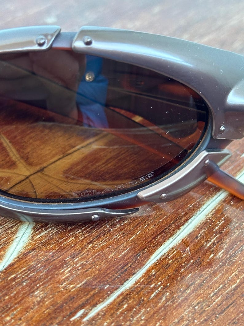 Puede incluir: Primer plano de unas gafas de sol marrones con montura gris oscuro. Las lentes son de color marr&oacute;n degradado. La montura tiene un dise&ntilde;o &uacute;nico con remaches visibles y una forma curva. Las gafas de sol est&aacute;n sobre una superficie de madera.