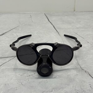 May include: Black sunglasses with round lenses and a unique bridge design. The sunglasses have a sleek, modern look with a dark frame and lenses. The arms curve around the ears, providing a secure fit. The sunglasses are designed for eye protection.