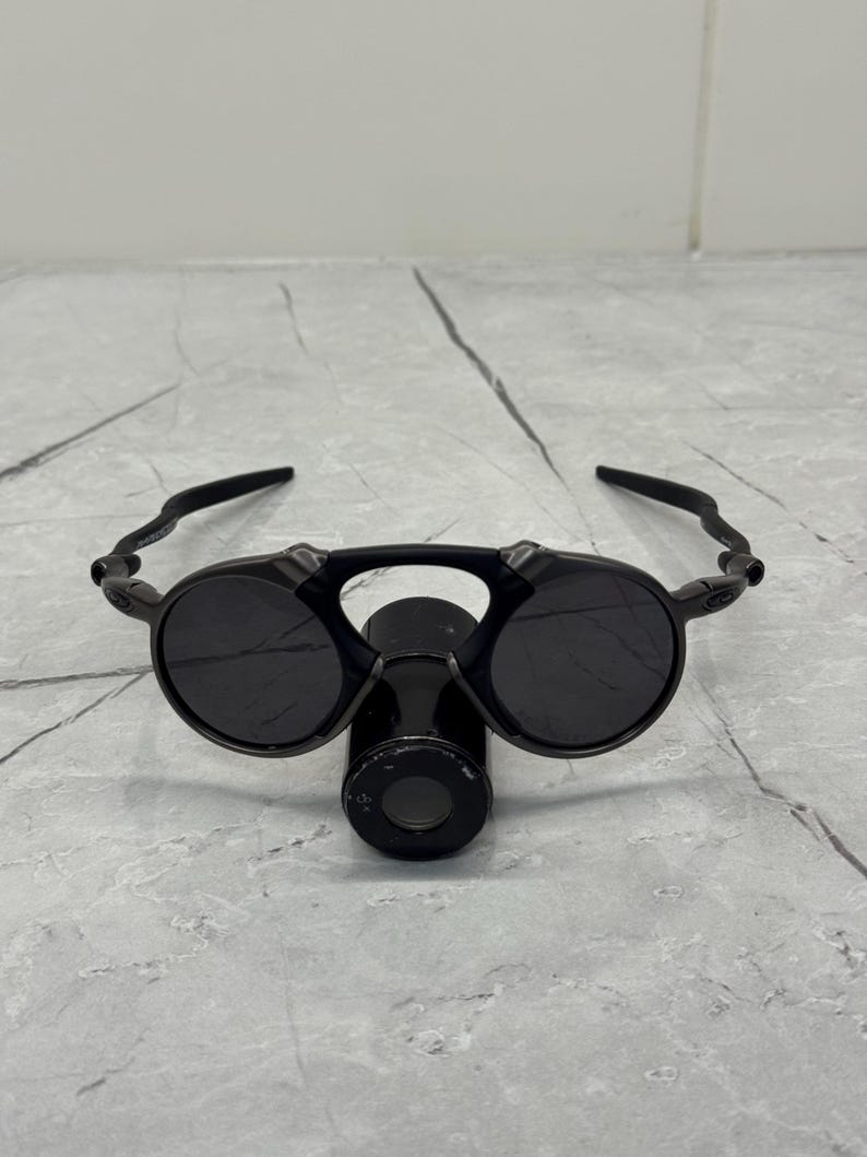 May include: Black sunglasses with round lenses and a unique bridge design. The sunglasses have a sleek, modern look with a dark frame and lenses. The arms curve around the ears, providing a secure fit. The sunglasses are designed for eye protection.