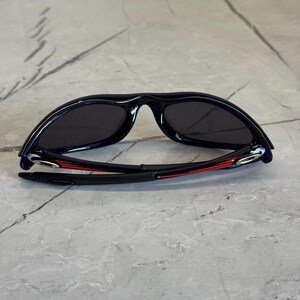 K&ouml;nnte beinhalten: Schwarze Sonnenbrille mit geschwungenem Rahmen und dunklen Gl&auml;sern. Die B&uuml;gel sind schwarz mit roten Akzenten. Die Sonnenbrille liegt auf einer grauen Oberfl&auml;che.