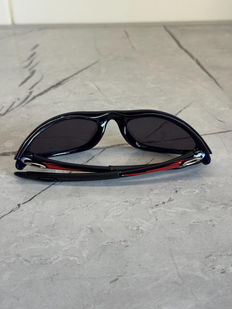 K&ouml;nnte beinhalten: Schwarze Sonnenbrille mit geschwungenem Rahmen und dunklen Gl&auml;sern. Die B&uuml;gel sind schwarz mit roten Akzenten. Die Sonnenbrille liegt auf einer grauen Oberfl&auml;che.