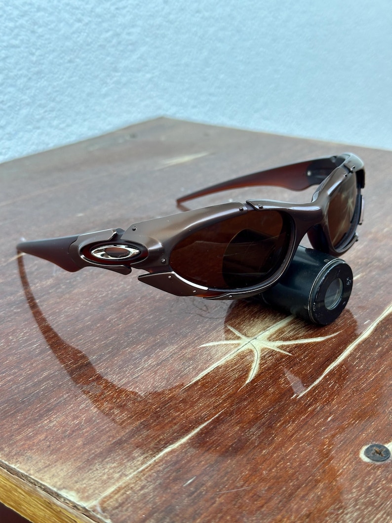 Puede incluir: Gafas de sol Oakley marrones con tinte oscuro, sobre una superficie de madera. Las gafas tienen un dise&ntilde;o deportivo con montura curvada y el logotipo de Oakley en el lateral.