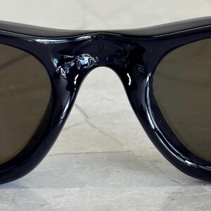 May include: Close-up of dark navy sunglasses with brown lenses. The bridge of the sunglasses has the letters "V" and "N" etched into the frame. The sunglasses are on a light gray surface.