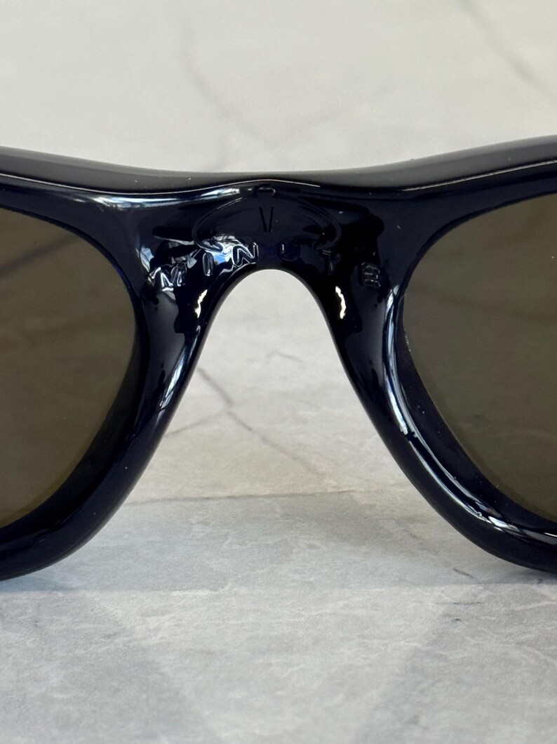 May include: Close-up of dark navy sunglasses with brown lenses. The bridge of the sunglasses has the letters "V" and "N" etched into the frame. The sunglasses are on a light gray surface.