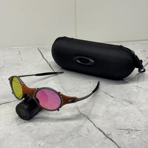 以下が含まれることがあります： 黒いフレーム、茶色の革のアクセント、楕円形のレンズのサングラス。 片方のレンズは黄色、もう片方はピンクです。 ジッパーとブランドロゴが付いた黒いケースも写っています。