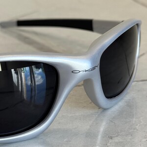 May include: Silver-framed sunglasses with dark lenses. The sunglasses have a sleek, curved design and the brand name "Oakley" is visible on the frame. The lenses reflect the surrounding environment.