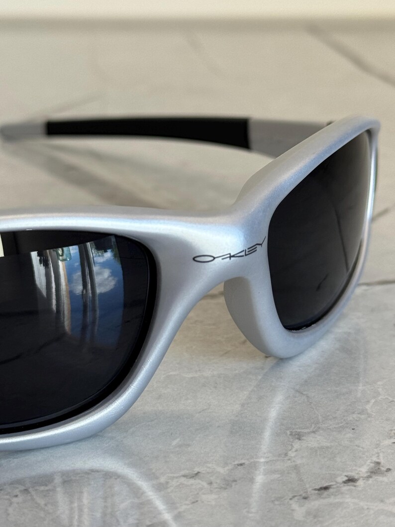 May include: Silver-framed sunglasses with dark lenses. The sunglasses have a sleek, curved design and the brand name "Oakley" is visible on the frame. The lenses reflect the surrounding environment.