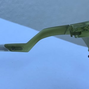 May include: Close-up of a pair of green-tinted eyeglasses. The frame is a light green color, with a curved arm and a hinge visible. The arm has text and numbers printed on it. The background is a light blue color.