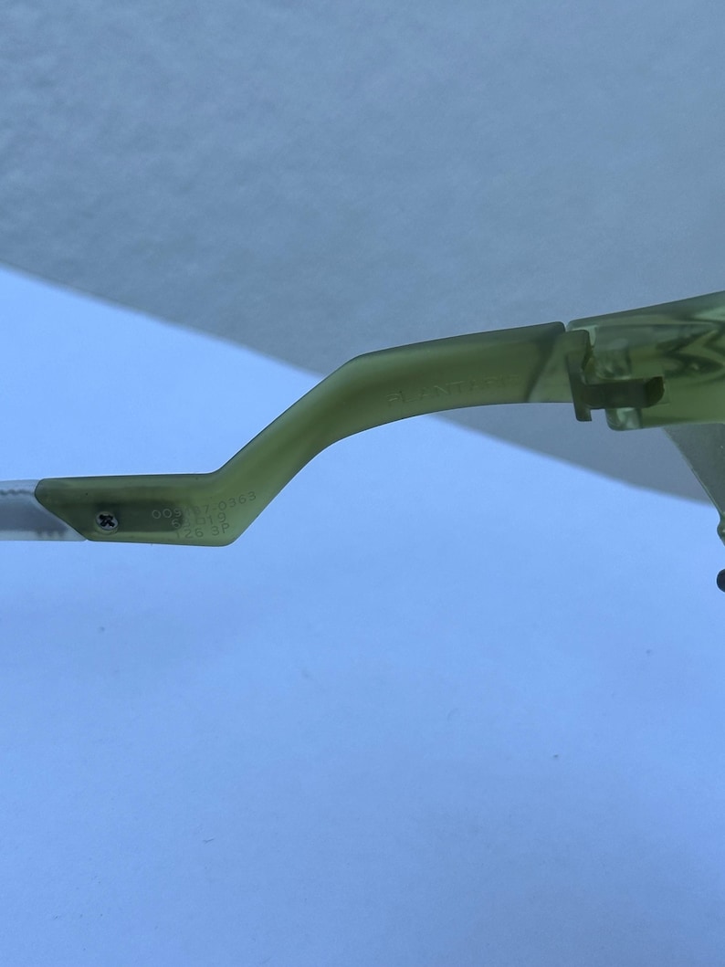 May include: Close-up of a pair of green-tinted eyeglasses. The frame is a light green color, with a curved arm and a hinge visible. The arm has text and numbers printed on it. The background is a light blue color.
