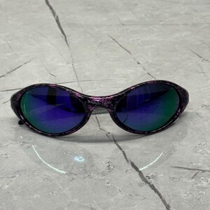 May include: Oval sunglasses with a black and purple speckled frame and mirrored lenses. The lenses reflect blue and green. The text "BRAIN DEAD" is printed on the bridge. A stylish accessory.