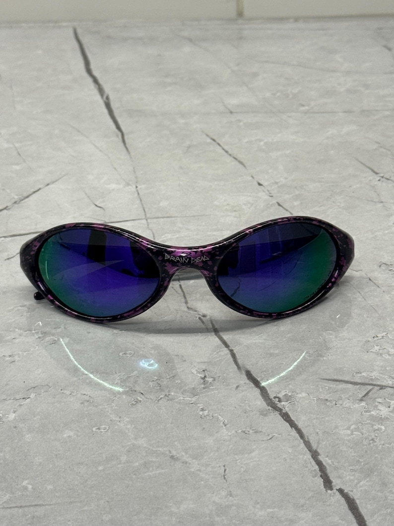 May include: Oval sunglasses with a black and purple speckled frame and mirrored lenses. The lenses reflect blue and green. The text "BRAIN DEAD" is printed on the bridge. A stylish accessory.