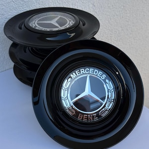 May include: A stack of four black hubcaps with a glossy finish. Each hubcap features a silver Mercedes-Benz logo in the center. The logo is a three-pointed star within a circular emblem, with the words "MERCEDES BENZ" around it.