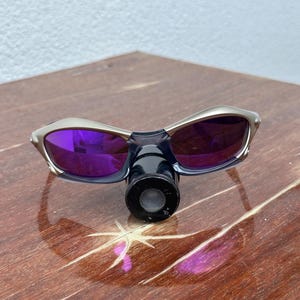 May include: Silver sunglasses with purple lenses and a black lens cap. The sunglasses are sitting on a wooden surface.