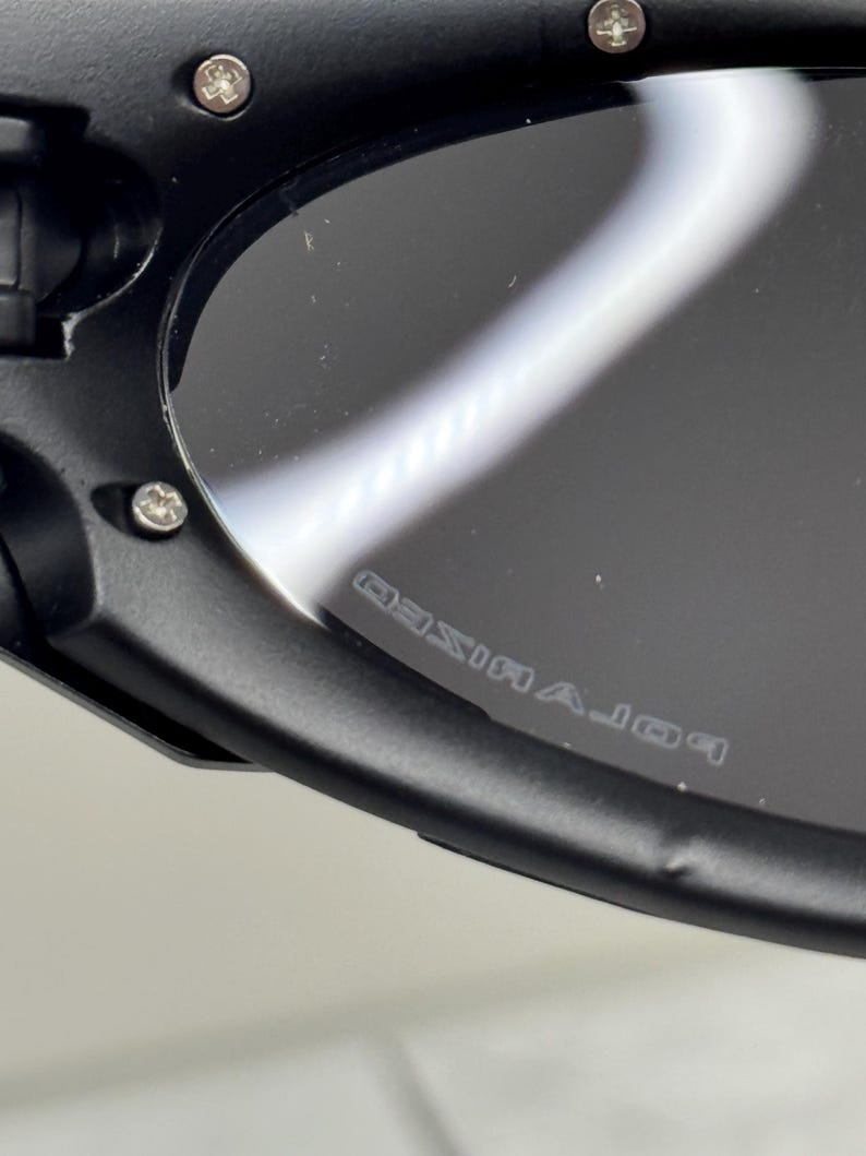 May include: Close-up of black sunglasses with polarized lenses. The frame is black and has silver rivets. The lenses are dark and reflect light. The word "POLARIZED" is printed on the lens.