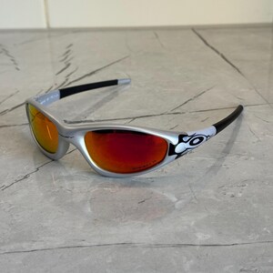 May include: Silver-framed sunglasses with red-tinted lenses. The temples are black with a white and black graphic near the hinges. The sunglasses are on a gray marble surface.