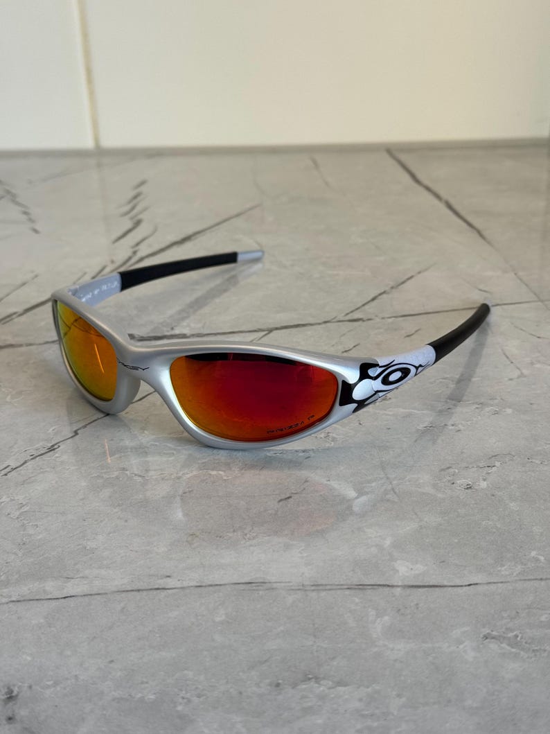 May include: Silver-framed sunglasses with red-tinted lenses. The temples are black with a white and black graphic near the hinges. The sunglasses are on a gray marble surface.