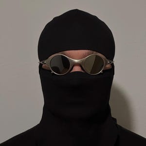 May include: Silver-framed sunglasses with round lenses are worn over a black face covering and a black cap. The sunglasses have a metallic finish and are the focal point of the image. The background is a plain, light gray wall.