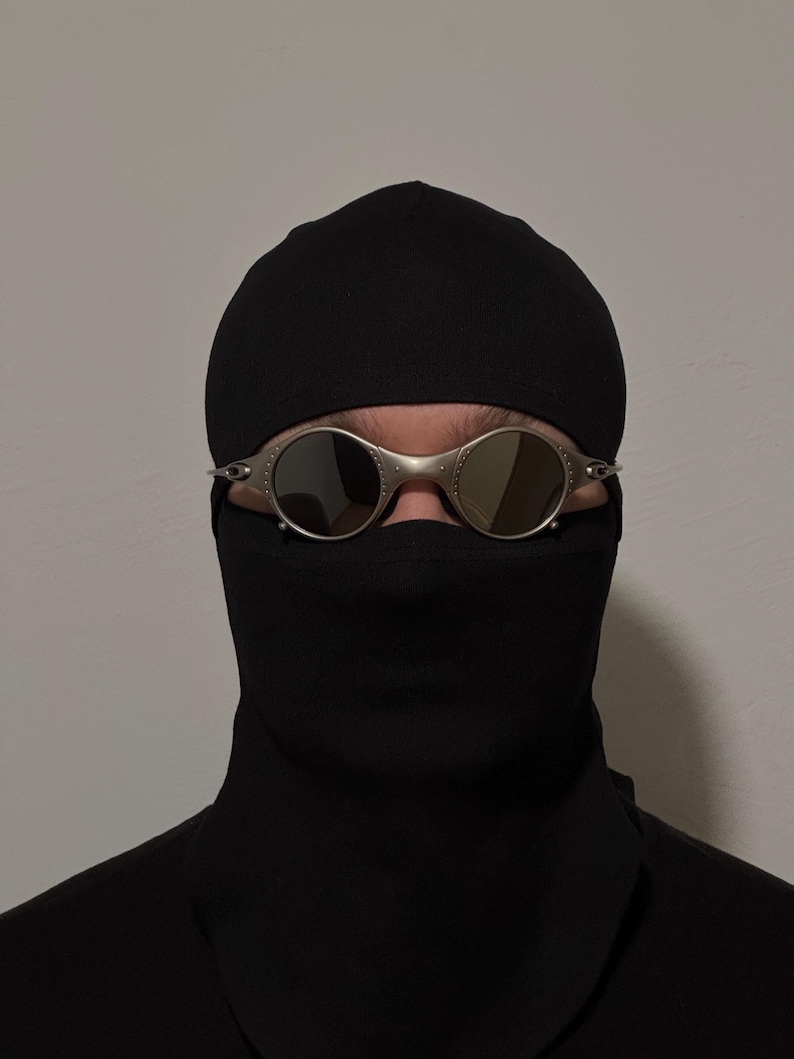 May include: Silver-framed sunglasses with round lenses are worn over a black face covering and a black cap. The sunglasses have a metallic finish and are the focal point of the image. The background is a plain, light gray wall.