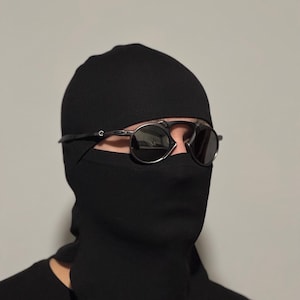 May include: A black balaclava with a face covering, worn with round, dark-tinted sunglasses. The sunglasses have a thin, silver frame. The balaclava covers the head and neck, leaving only the eyes visible.