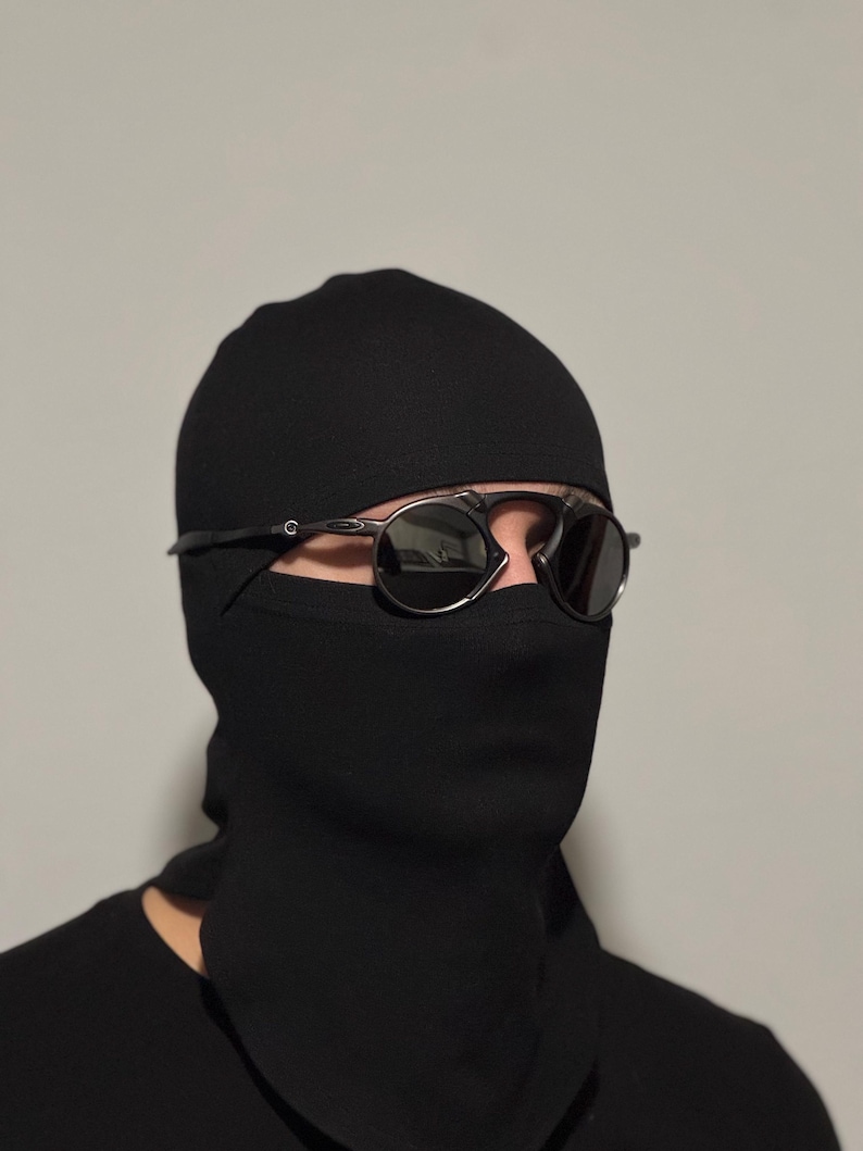 May include: A black balaclava with a face covering, worn with round, dark-tinted sunglasses. The sunglasses have a thin, silver frame. The balaclava covers the head and neck, leaving only the eyes visible.